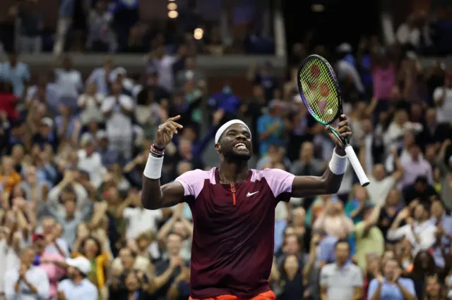 Frances Tiafoe celebrates with a big smile and both hands raised to the crowd with the fans celebrating in the stands out of focus in the background behind him