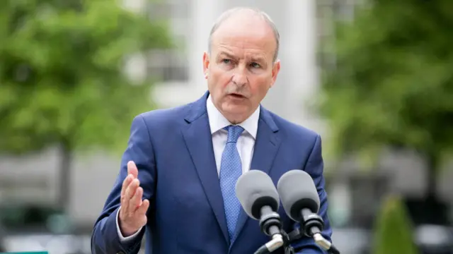 Tánaiste (Irish deputy prime minister) Micheál Martin standing infront of two microphones. The background is blurred.