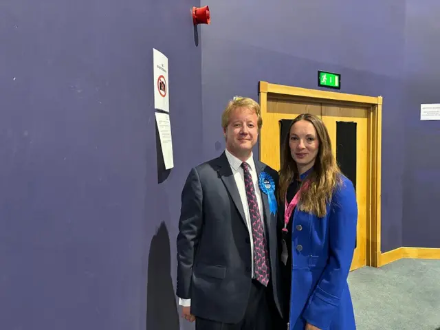 Paul Bristow wearing a navy suit and tie with a blue rosette has his arm around a woman in a blue jacket.