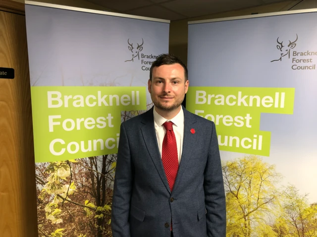 Peter Swallow is wearing a dark suit with a red tie in front of Bracknell Forest Council signs