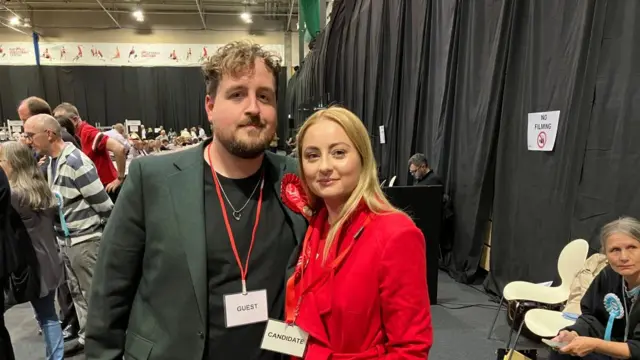 Gen Kitchen with long blond hair and in a red jacket at the count standing next to a man