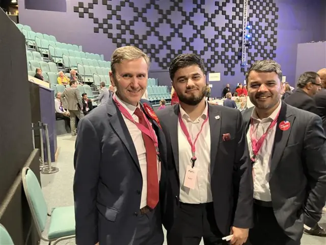 A smiling Andrew Pakes alongside two other men, all wearing suits and pink lanyards and stood in a convention hall.