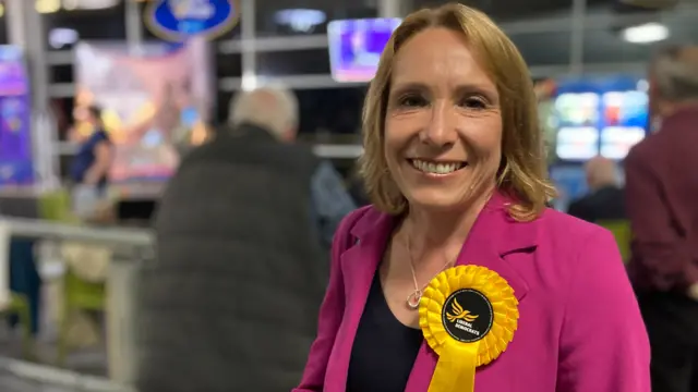 A smiling Helen Morgan at the count, wearing a pink jacket and Lib Dem rosette