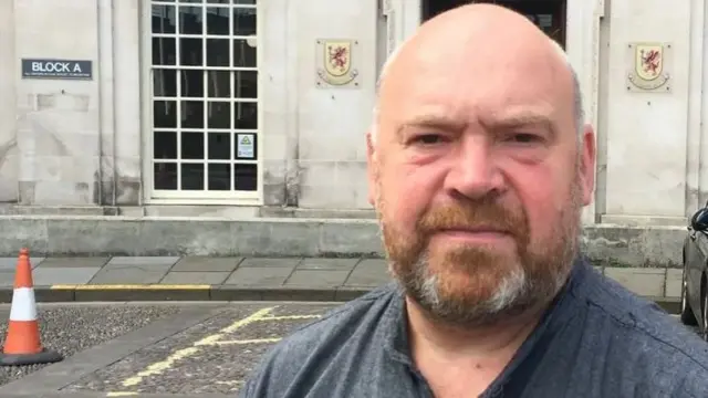 Head and shoulders picture of Bill Revans wearing a grey shirt, standing in front of the Somerset Council building in Taunton