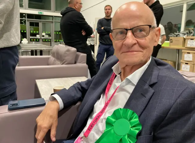 A man with glasses and a blue suit and a green rosette