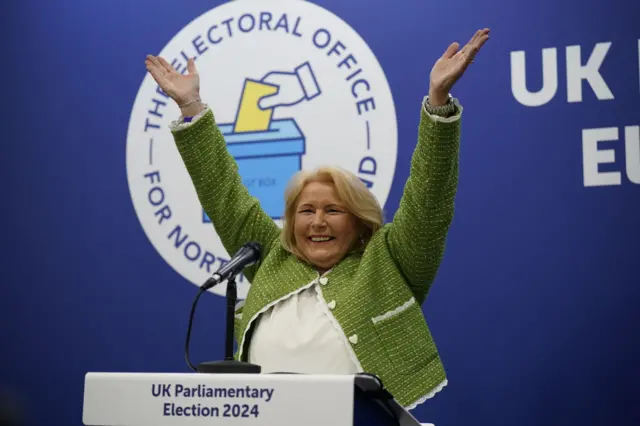 Blonde woman with her arms up in a green jacket and white shirt