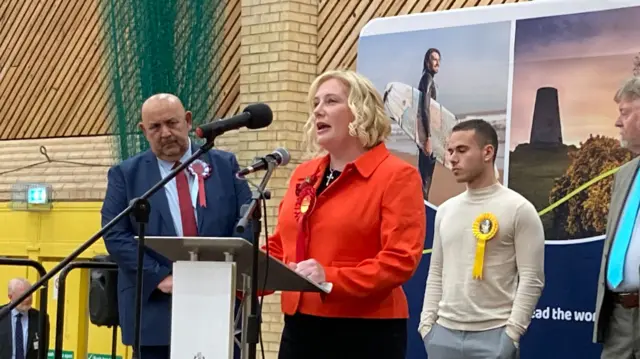 A woman in a red jacket makes a speech on a stage