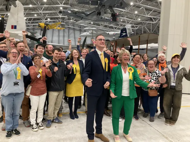 Pippa Heylings in a green suit cheering, standing alongside a male in a suit and a number of cheering supporters behind her.