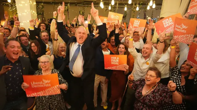 Ed Davey jumping in the air with hands raised amid sea of Lib Dem activists holding orange placards