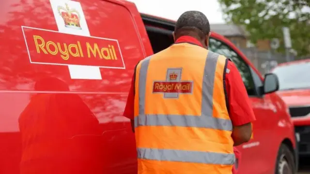 Royal Mail worker next to a van, getting something out the back. His back is turned to the camera