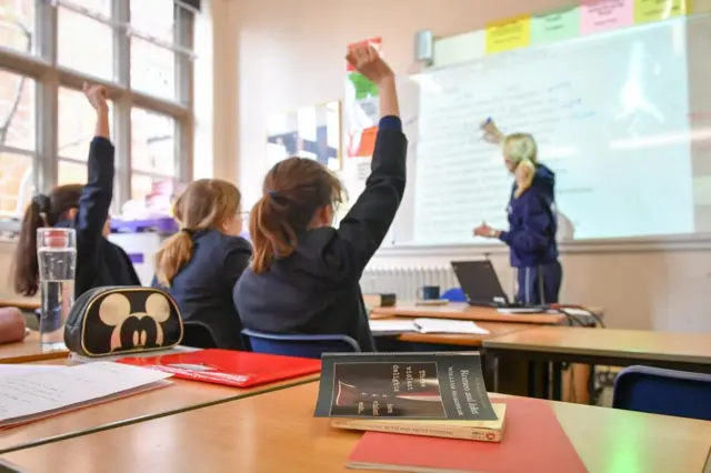 Interior shot of a classroom with students putting their hands up (viewed from behind) and a teacher writing on a whiteboard in the background