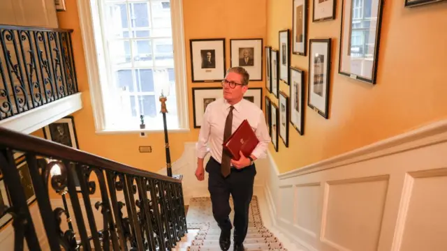 M Keir Starmer walking past portraits of previous UK Prime Ministers on the stairs inside Number Ten Downing Street