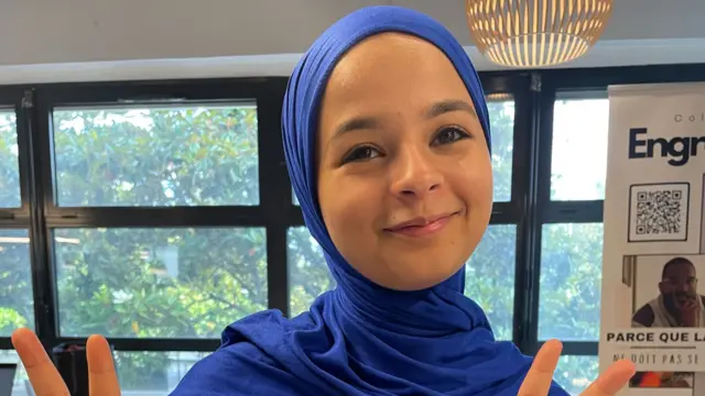 Sarah Bennani wearing a blue hijab, smiling holding up a peace sign.