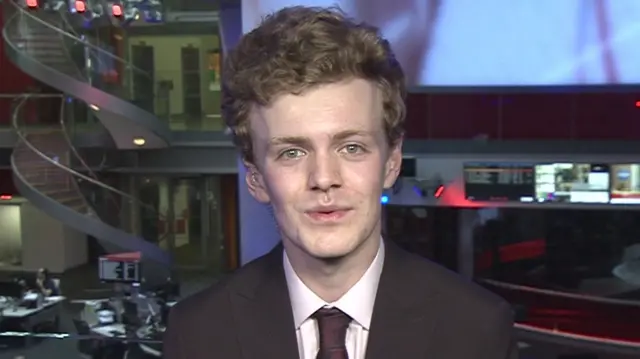 Sam Carling in dark suit looking into camera with BBC newsroom behind him