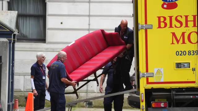 Two men carry a bright red striped sofa up a ramp and into the back of a bright yellow removal van