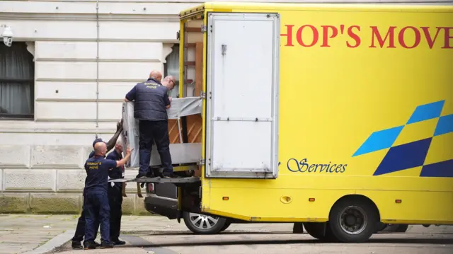 Piano covered in white sheet being carried into the back of a bright yellow removal van by a group of men