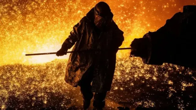 Sparks fly as a worker in a heat suit and and hood changes the nozzle on a clay gun in a blast furnace in Port Talbot