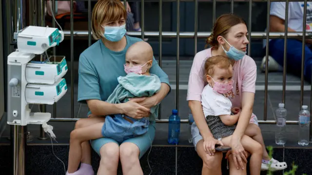 Two women wearing face masks holding a child each near the hospital.