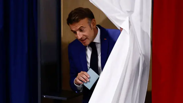 Macron holds his voting card as he walks through a white curtain after voting in the first round of the parliamentary elections