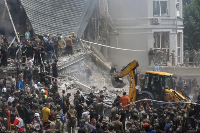 Dozens of rescue workers at the site of the missile hit on the hospital, with a JCB digger removing reubble nearby