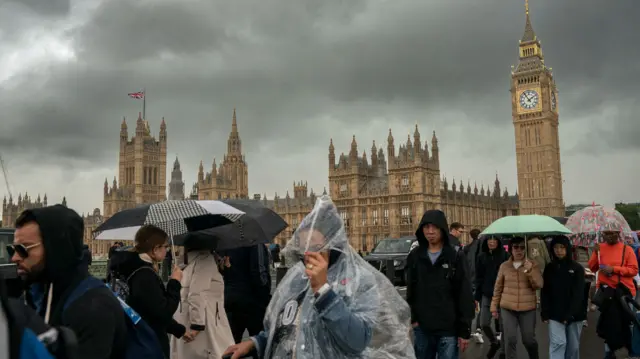 People with umbrellas and ponchos walk through heavy rain with Westminster in the backgroun