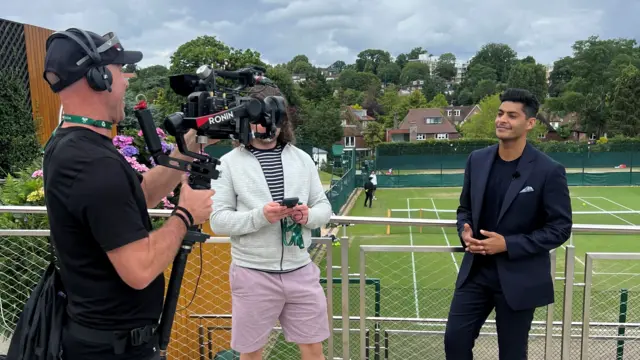 Cameraman with a camera filming a presenter in front of a tennis court