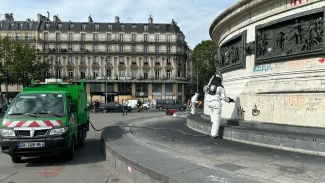 Person in white overalls cleans graffiti off tall white statue in middle of street, carrying a hose connected to a bright green vehicle