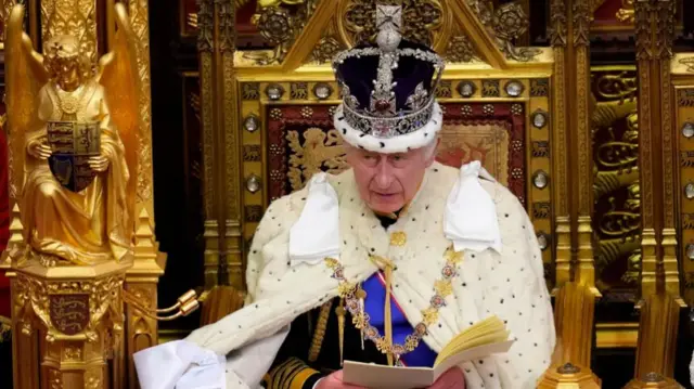 King Charles sits on a golden throne wearing the crown jewels, reading a speech