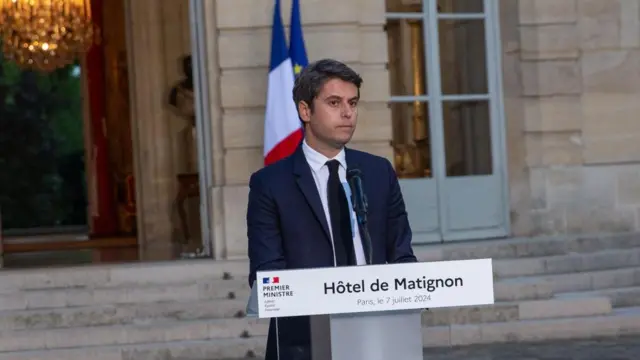 Gabriel Attal behind a lectern, he is front of a brick wall