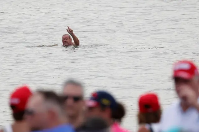 A man swimming in a pond, raising his hand, with a crowd in the foreground