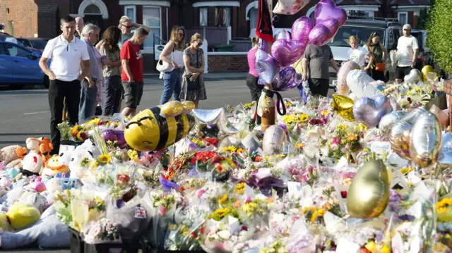 People stand in front of floral tributes and balloons