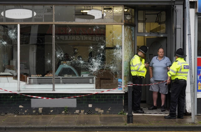 A man speaks to two police officers at the door of a smashed butchers shop