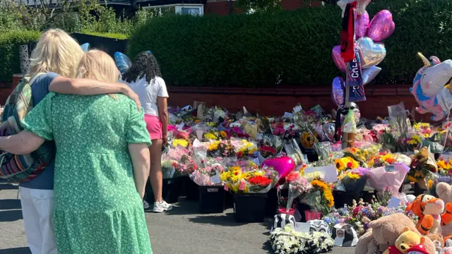 A woman stands with her arm around another next to a tribute of flowers and toys in Southport