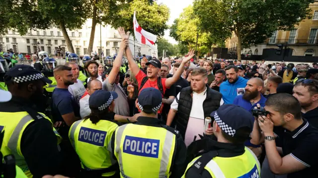 Protesters stand before a police line in central London, with an England flag waved in the background