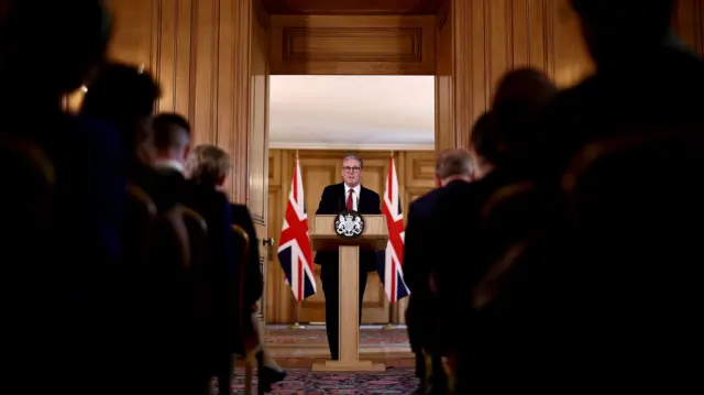 Sir Keir Starmer stands in front of a lectern at the end of an aisle with people on chairs either side of the aisle