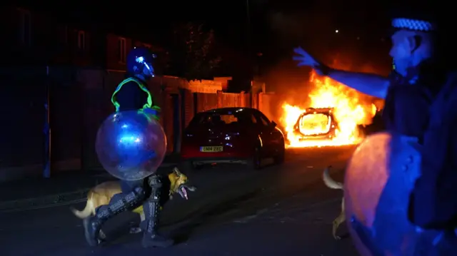 A police car burns and a police officer in riot gear walks with a police dog in Hartlepool