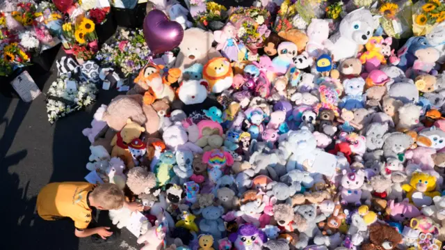 A boy bends down near a big pile of soft toys and teddies covering the pavement