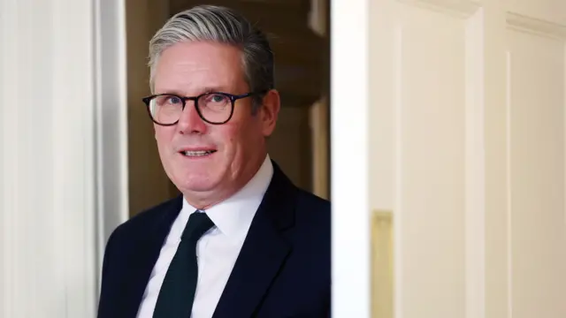 Sir Keir Starmer smiles while walking through a door. He is wearing a black suit and tie