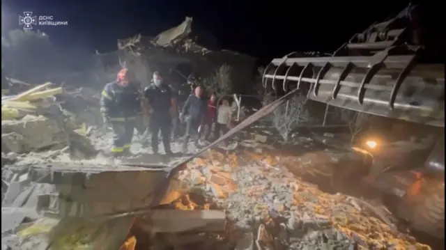 Men, one in protective gear, stand among rubble with the scene lit up by artificial light