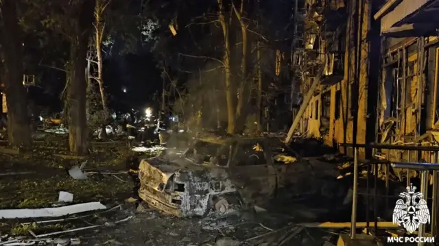 A burnt car stands among rubble and damaged buildings