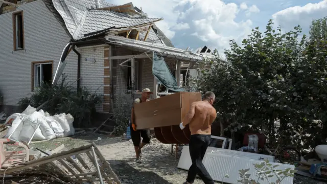 Two men lift a wardrobe against the backdrop of a damaged house