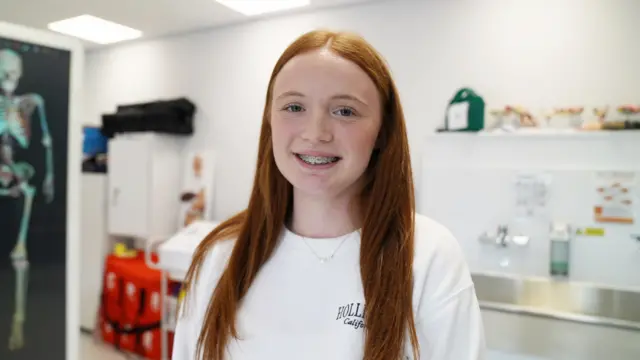 Katy is in a classroom with a sink and computer screen with a skeleton on in the background. She smiles at the camera and is wearing a white top and has long red hair