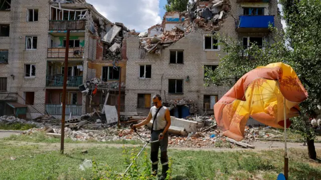 A man mows the grass near a residential building, which was heavily damaged in the course of Russia-Ukraine conflict