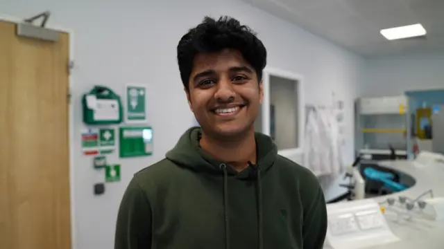 Eshan stands in a classroom and smiles at the camera. He has short, black hair and is wearing a green hoodie