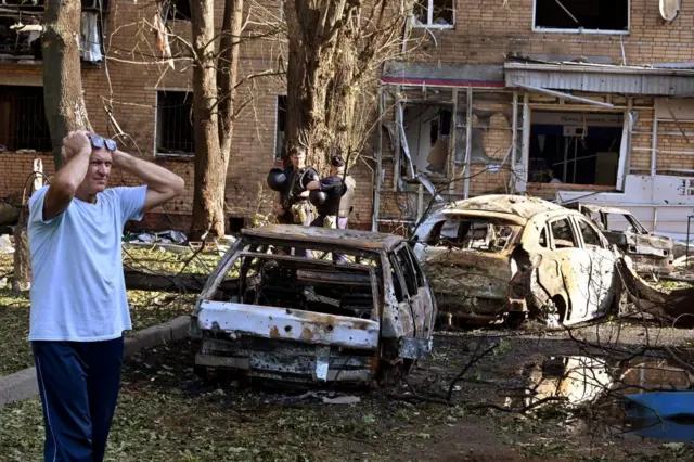 A man with his hands on his head stood in front of two burned out cars