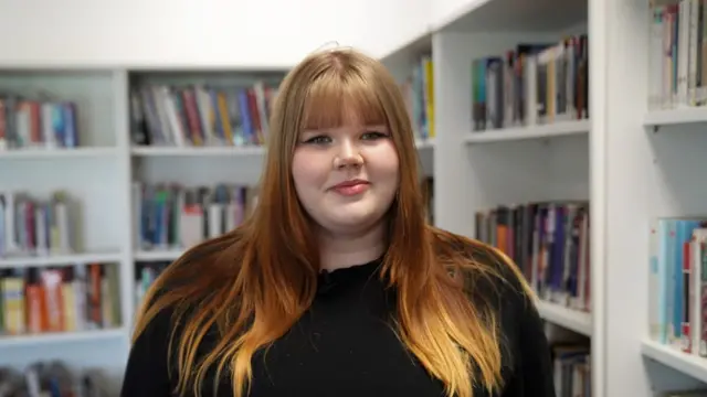 Ruby is looking at the camera and smiling. There are bookshelves in the background. She’s wearing a black top and has long red hair.