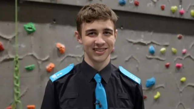 Benji is stood in front of a climbing wall smiling at the camera, wearing a black shirt and a blue tie