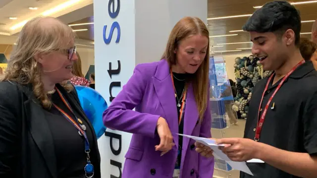 Two college staff members are on the left of a male student who is smiling at his results. The staff member in the middle is wearing a bright purple blazer.