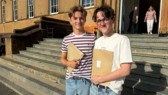 Two male students in jeans and T-shirts stand on some outdoor steps smiling to the camera with their result envelopes