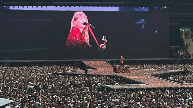 Taylor Swift playing guitar on stage in Wembley Stadium with thousands of fans